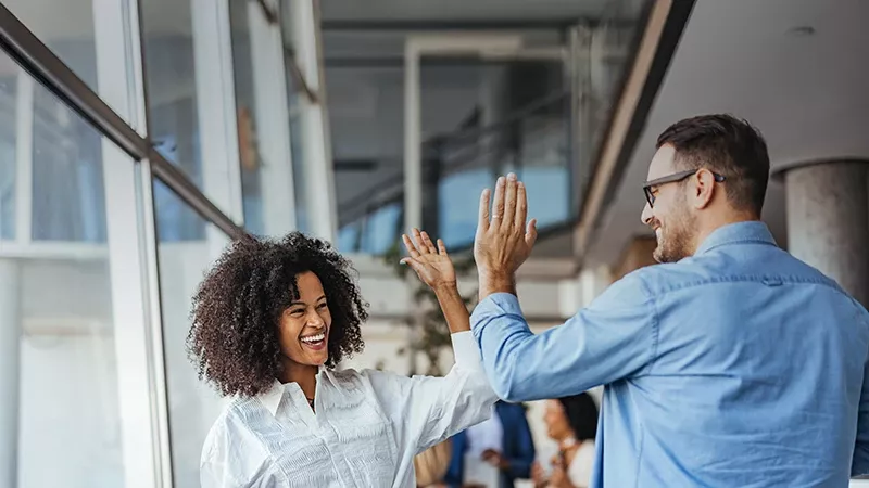 A stunning image displays a joyful embrace in a modern office, where a man in blue and a woman with curly hair beam with happiness. The glass wall adds a sleek touch, while the blurred background hints at a lively, bustling environment. This image captures camaraderie in a visually captivating moment.