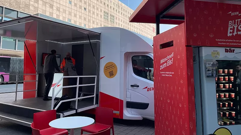 A mobile ice cream truck with a serving area and vending machine set up in an urban location, featuring red seating and signage promoting ice cream. Two people are entering the truck.