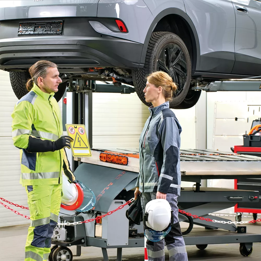 Two workers in safety gear discuss under a raised electric vehicle. One holds a hazard sign. Both wear helmets and high-visibility clothing. The garage has tools and machinery visible.