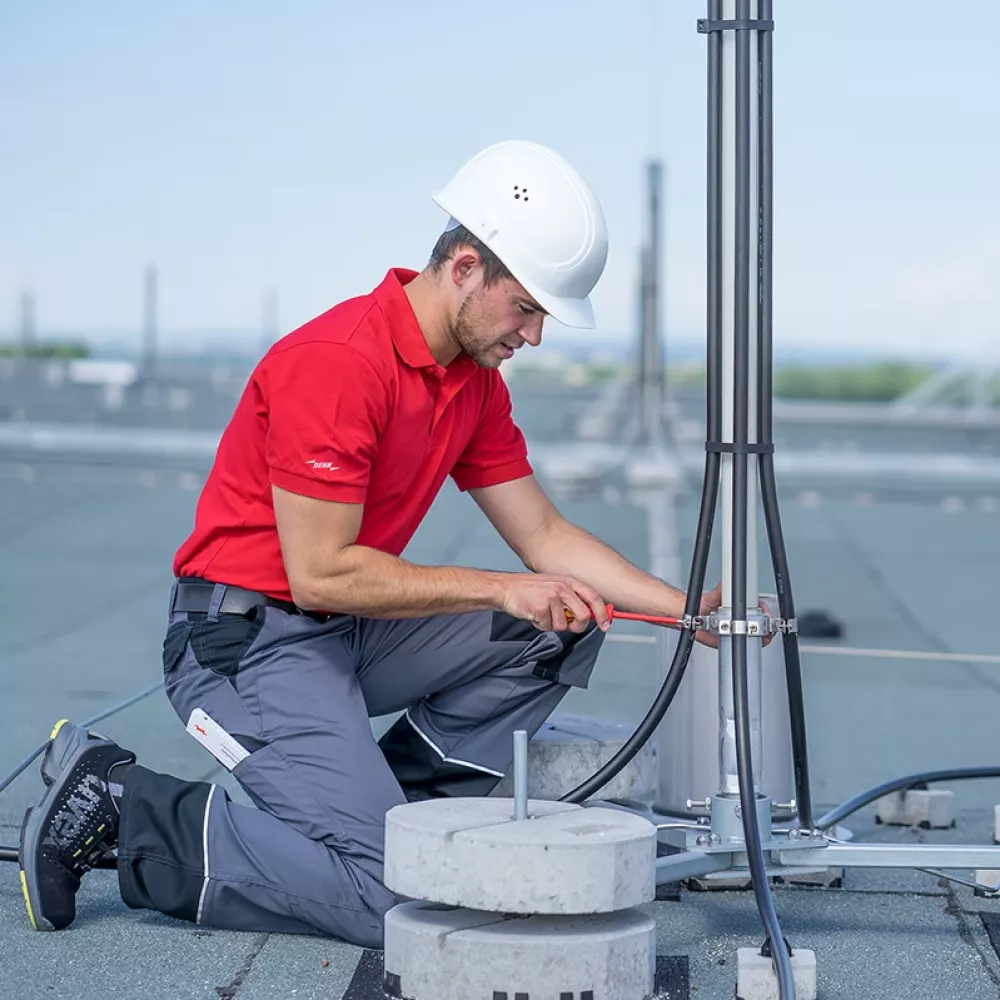 Technician in red shirt and white hard hat installing grounding cables on a rooftop lightning protection mast.